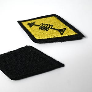 K&ouml;nnte beinhalten: Zwei rautenf&ouml;rmige Aufn&auml;her. Einer ist schwarz, der andere ist gelb mit einem schwarzen Pfeil- und Spulendesign. Die Aufn&auml;her sind wahrscheinlich f&uuml;r Kleidung oder Accessoires gedacht und bieten eine M&ouml;glichkeit, Artikel zu personalisieren.