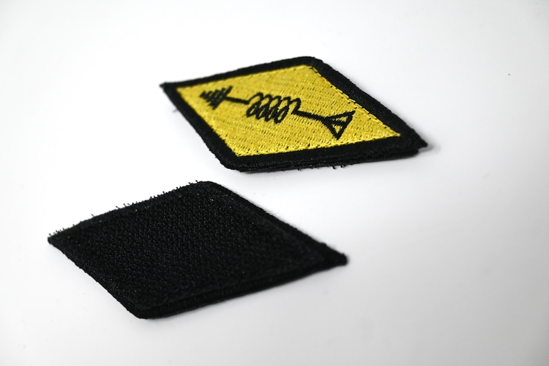 K&ouml;nnte beinhalten: Zwei rautenf&ouml;rmige Aufn&auml;her. Einer ist schwarz, der andere ist gelb mit einem schwarzen Pfeil- und Spulendesign. Die Aufn&auml;her sind wahrscheinlich f&uuml;r Kleidung oder Accessoires gedacht und bieten eine M&ouml;glichkeit, Artikel zu personalisieren.