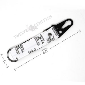 May include: A white fabric keychain with a black metal clasp and a ring. The fabric has the text "Desiccant Silica Gel Throw Away Do Not Eat" printed on it. The keychain is approximately 6.5 inches long.