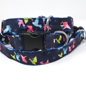 May include: A blue dog collar with a colorful origami bird pattern. The collar has a black plastic buckle and silver metal hardware.