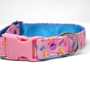 May include: Pink dog collar with a sprinkle pattern, featuring a pink plastic buckle and a silver metal D-ring. The collar's interior is a bright blue, and it has a purple jewel accent.