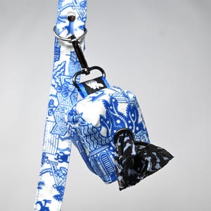 May include: Blue and white patterned dog waste bag dispenser with a black bag attached. The dispenser has a blue strap with a silver clip.