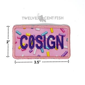 May include: A rectangular embroidered patch with a pink background featuring a colorful sprinkle pattern and the word COSIGN in purple capital letters. The patch measures 2 inches by 3.5 inches as indicated by the visible measurement markings.