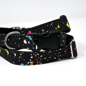May include: Black dog collar with a colorful paint splatter pattern. The collar has a silver D-ring and a black plastic buckle.