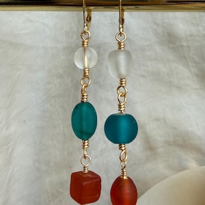 May include: A pair of dangle earrings featuring gold-tone wire and hardware. Each earring consists of three stacked frosted glass beads in clear, teal, and orange. The left earring has a cube-shaped orange bead, while the right has an oval orange bead.