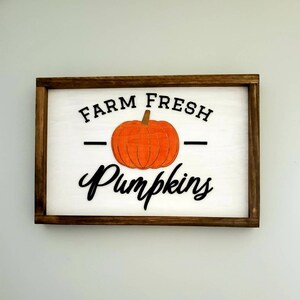 Puede incluir: Un letrero de madera con marco marr&oacute;n y fondo blanco. El letrero dice "Farm Fresh Pumpkins" con una ilustraci&oacute;n de calabaza negra y naranja.