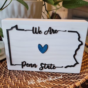 May include: White wooden sign with a black outline of the state of Pennsylvania. The sign says "We Are Penn State" with a blue heart in the centre.