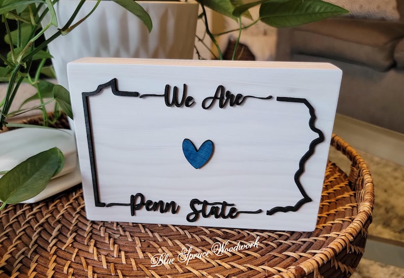 May include: White wooden sign with a black outline of the state of Pennsylvania. The sign says "We Are Penn State" with a blue heart in the centre.