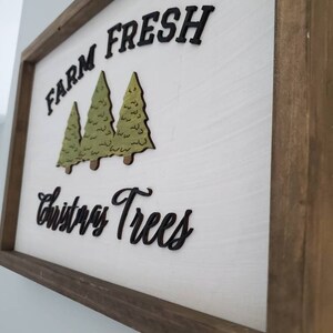 Puede incluir: Un letrero de madera con fondo blanco y marco marr&oacute;n. El letrero dice "Farm Fresh Christmas Trees" en letras negras. Hay tres &aacute;rboles de Navidad verdes recortados en madera y pegados al letrero.