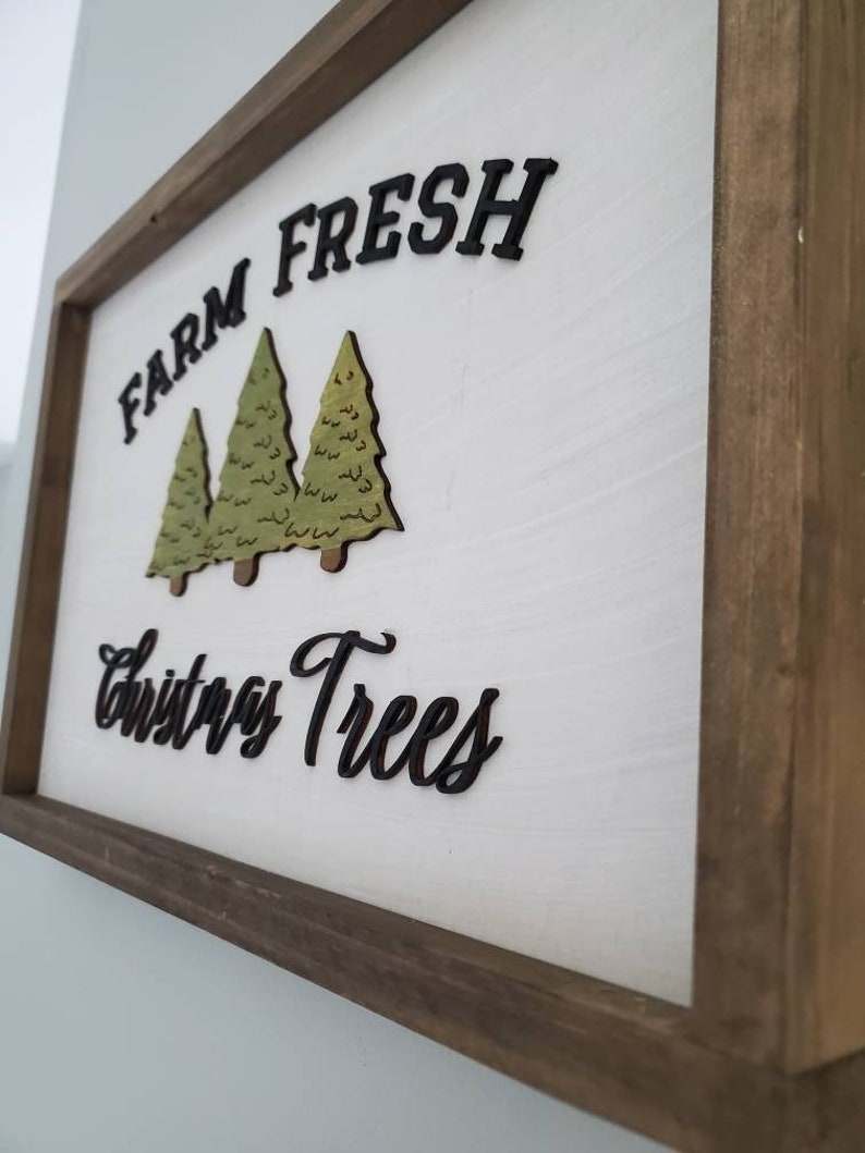 Puede incluir: Un letrero de madera con fondo blanco y marco marr&oacute;n. El letrero dice "Farm Fresh Christmas Trees" en letras negras. Hay tres &aacute;rboles de Navidad verdes recortados en madera y pegados al letrero.