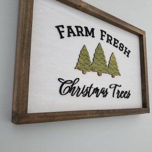 Puede incluir: Un letrero de madera con fondo blanco y marco marr&oacute;n. El letrero dice "Farm Fresh Christmas Trees" en letras negras. Hay tres &aacute;rboles de Navidad verdes recortados en madera y pegados al letrero.