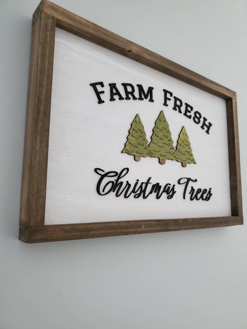 Puede incluir: Un letrero de madera con fondo blanco y marco marr&oacute;n. El letrero dice "Farm Fresh Christmas Trees" en letras negras. Hay tres &aacute;rboles de Navidad verdes recortados en madera y pegados al letrero.