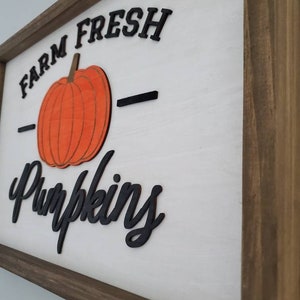 Puede incluir: Un letrero de madera con marco marr&oacute;n y fondo blanco. El letrero dice "Farm Fresh Pumpkins" en letras negras con una gran calabaza naranja en el centro.