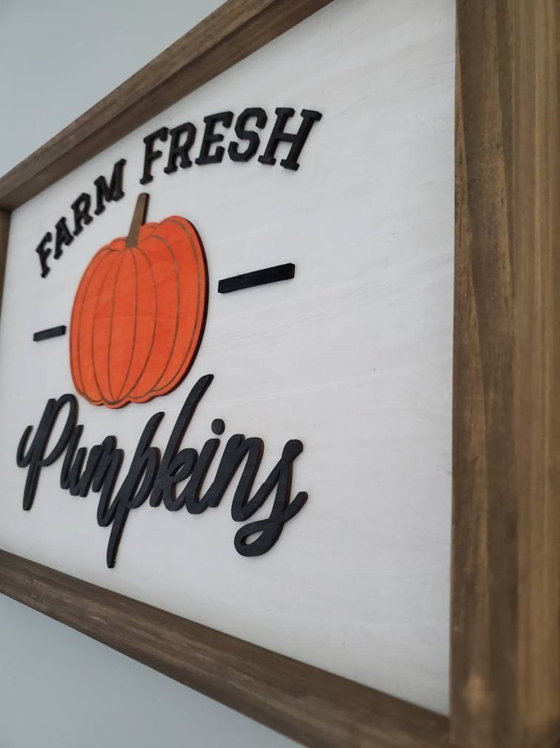 Puede incluir: Un letrero de madera con marco marr&oacute;n y fondo blanco. El letrero dice "Farm Fresh Pumpkins" en letras negras con una gran calabaza naranja en el centro.