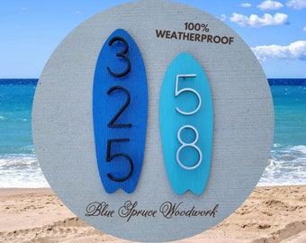 May include: Two blue surfboard-shaped house number signs with black numbers. The sign on the left has the numbers 325 and the sign on the right has the numbers 58. The signs are weatherproof and have the text "100% WEATHERPROOF" and "Blue Spruce Woodwork" on them.
