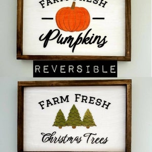 Puede incluir: Un letrero de madera reversible con fondo blanco y marco marr&oacute;n. Un lado presenta el texto "Farm Fresh Pumpkins" con una ilustraci&oacute;n de calabaza negra. El otro lado presenta el texto "Farm Fresh Christmas Trees" con tres ilustraciones de &aacute;rboles de Navidad verdes.