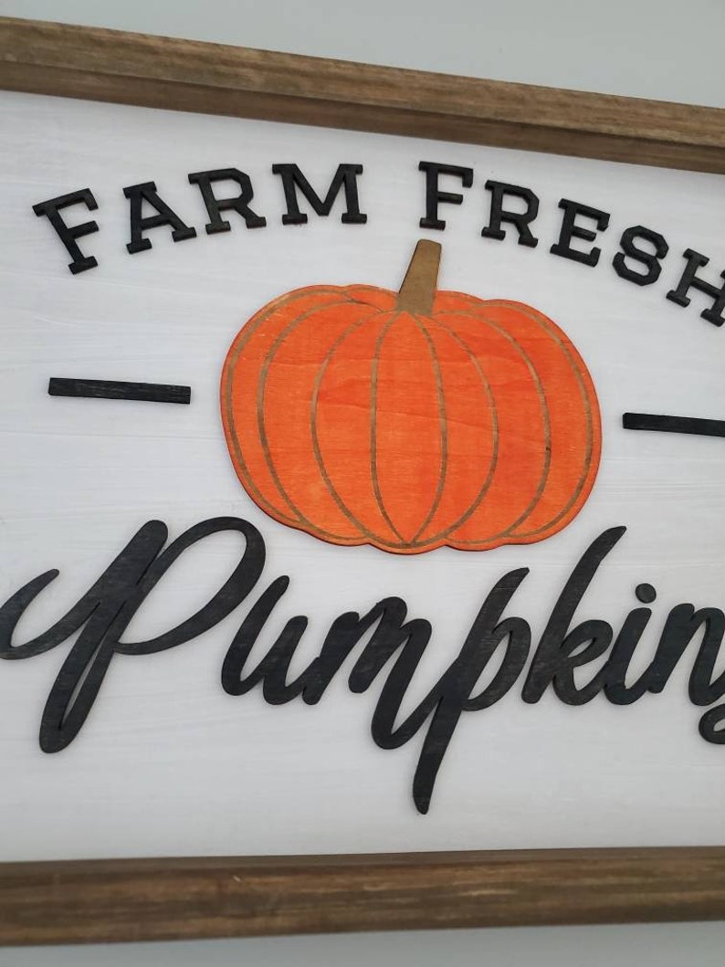 Puede incluir: Un letrero de madera blanco con marco marr&oacute;n, con el texto "FARM FRESH PUMPKINS" en letras negras. Una gran calabaza naranja con un tallo marr&oacute;n est&aacute; centrada en el letrero.