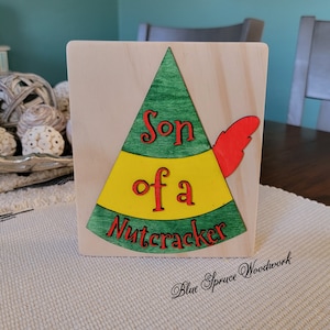 May include: Wooden sign with a green, yellow, and red elf hat design. The sign reads "Son of a Nutcracker".
