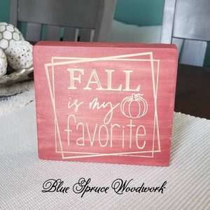 May include: A red wooden sign with the text "FALL is my favorite" in white lettering. A small pumpkin is drawn next to the word "my". The sign is distressed and has a rustic look.
