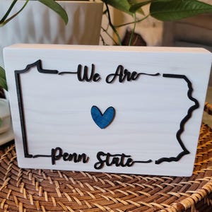 May include: White wooden sign with a black outline of the state of Pennsylvania. The sign says "We Are Penn State" with a blue heart in the centre.