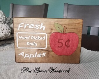May include: Wooden sign with the text "Fresh Hand Picked Daily Apples" and a red apple with a green leaf and the text "5¢" on it. The sign is made by Blue Spruce Woodwork.