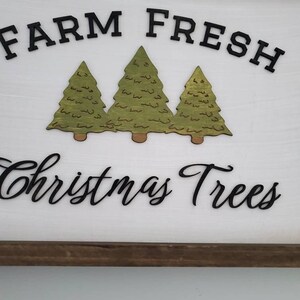 Puede incluir: Un letrero de madera blanco con un marco de madera marr&oacute;n. El letrero dice "Farm Fresh Christmas Trees" en letras negras. Hay tres &aacute;rboles de Navidad verdes recortados en madera y pegados al letrero.