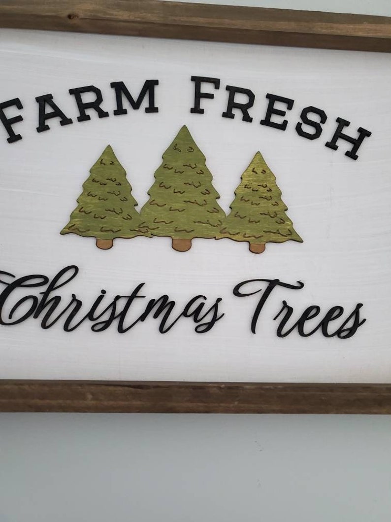 Puede incluir: Un letrero de madera blanco con un marco de madera marr&oacute;n. El letrero dice "Farm Fresh Christmas Trees" en letras negras. Hay tres &aacute;rboles de Navidad verdes recortados en madera y pegados al letrero.