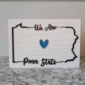 May include: Wooden sign with a black outline of the state of Pennsylvania. The sign reads "We Are Penn State" with a blue heart in the centre.