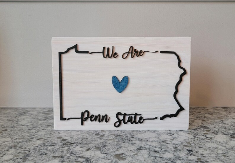 May include: Wooden sign with a black outline of the state of Pennsylvania. The sign reads "We Are Penn State" with a blue heart in the centre.