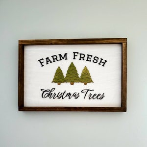 Puede incluir: Un letrero de madera blanco con marco marr&oacute;n dice "Farm Fresh Christmas Trees" en letras negras. Tres &aacute;rboles de Navidad verdes se representan en el centro del letrero.
