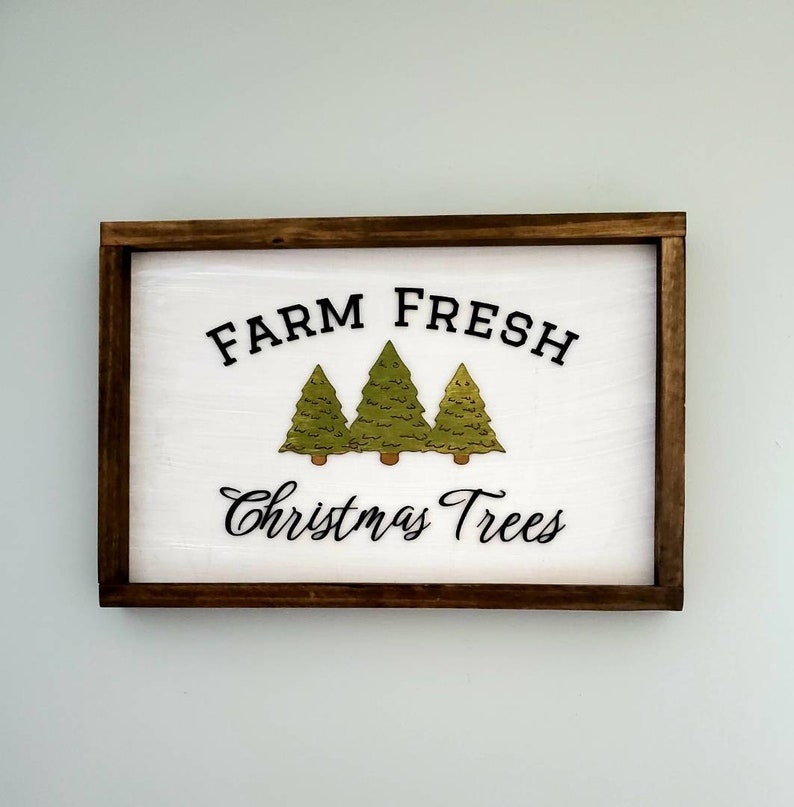Puede incluir: Un letrero de madera blanco con marco marr&oacute;n dice "Farm Fresh Christmas Trees" en letras negras. Tres &aacute;rboles de Navidad verdes se representan en el centro del letrero.