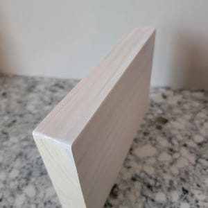 May include: A white wooden block with a smooth, painted surface. The block is rectangular and has a slightly rounded edge. The block is sitting on a light grey surface.