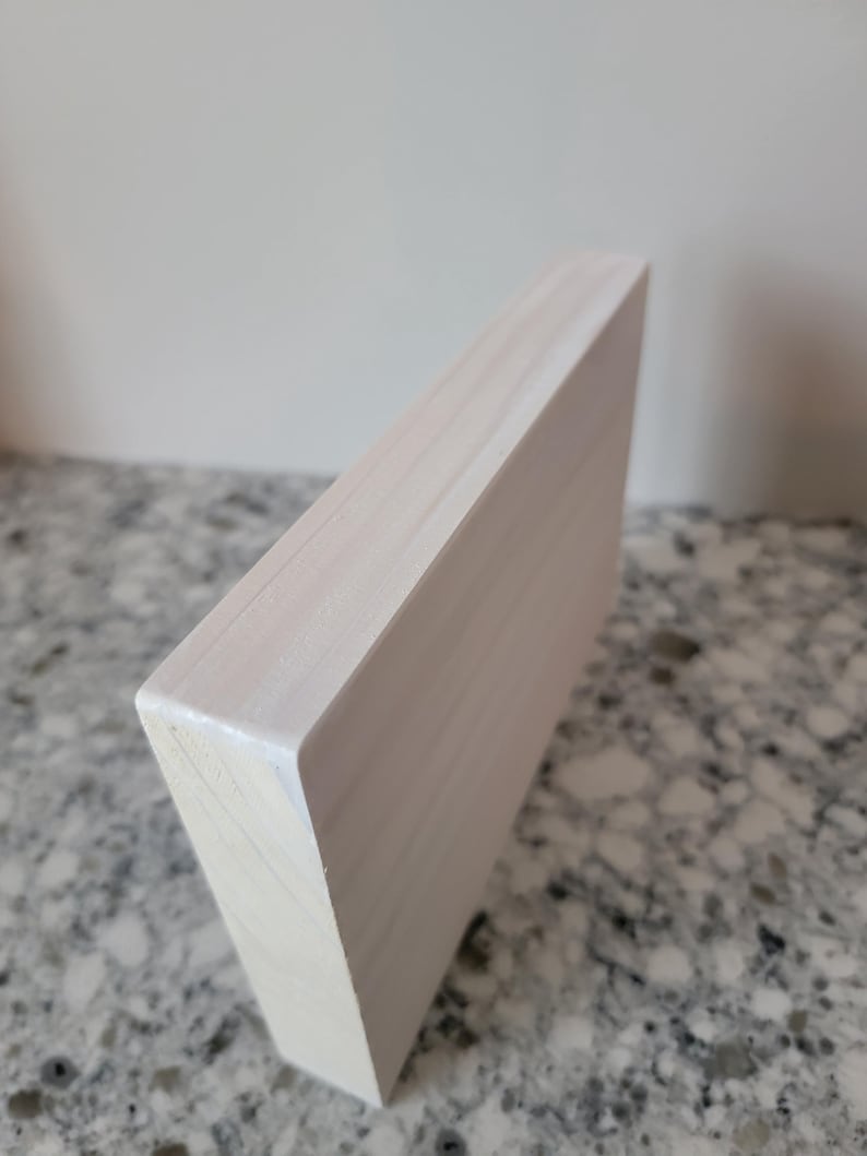 May include: A white wooden block with a smooth, painted surface. The block is rectangular and has a slightly rounded edge. The block is sitting on a light grey surface.