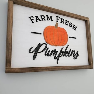 Puede incluir: Un letrero de madera con marco marr&oacute;n y fondo blanco. El letrero dice "FARM FRESH" con una calabaza negra en el medio y la palabra "Pumpkins" en una fuente cursiva negra.