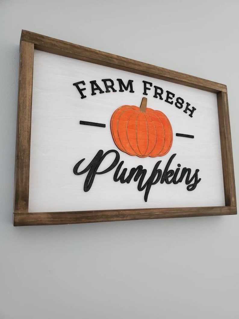 Puede incluir: Un letrero de madera con marco marr&oacute;n y fondo blanco. El letrero dice "FARM FRESH" con una calabaza negra en el medio y la palabra "Pumpkins" en una fuente cursiva negra.