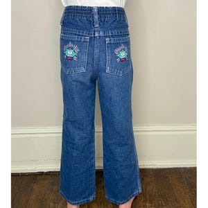 Könnte beinhalten: Blaue Jeans aus Denim mit gestickten Blumen und dem Text "Cherished Moments" auf den Gesäßtaschen.
