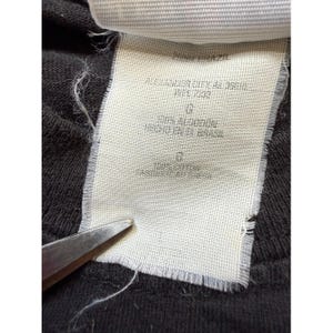 Pu&ograve; includere: Primo piano di un'etichetta di abbigliamento con testo. L'etichetta &egrave; bianca con testo nero ed &egrave; attaccata a un tessuto grigio scuro. Il testo include "ALEXANDER ZHER A5 35010" e "WPL 7232". L'etichetta dice anche "100% ALGODON HECHO EN BRASIL".