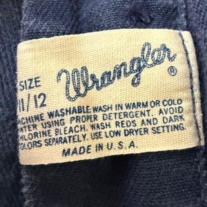 May include: Close-up of a Wrangler label on blue denim fabric. The label is cream-colored with blue text, displaying the brand name, size 11/12, and washing instructions. The label reads "Made in U.S.A."