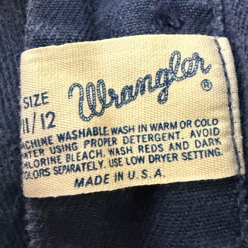 May include: Close-up of a Wrangler label on blue denim fabric. The label is cream-colored with blue text, displaying the brand name, size 11/12, and washing instructions. The label reads "Made in U.S.A."