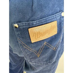 May include: Close-up of blue denim jeans with a back pocket. The pocket features a tan leather patch with the embossed word "Wrangler". The stitching is a contrasting gold color, and the jeans have silver rivets.