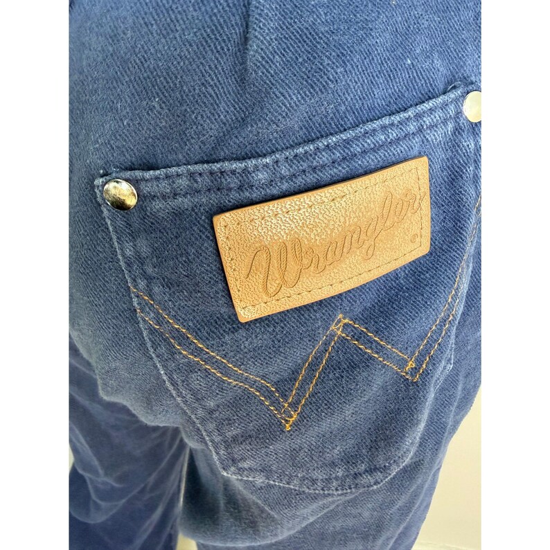 May include: Close-up of blue denim jeans with a back pocket. The pocket features a tan leather patch with the embossed word "Wrangler". The stitching is a contrasting gold color, and the jeans have silver rivets.