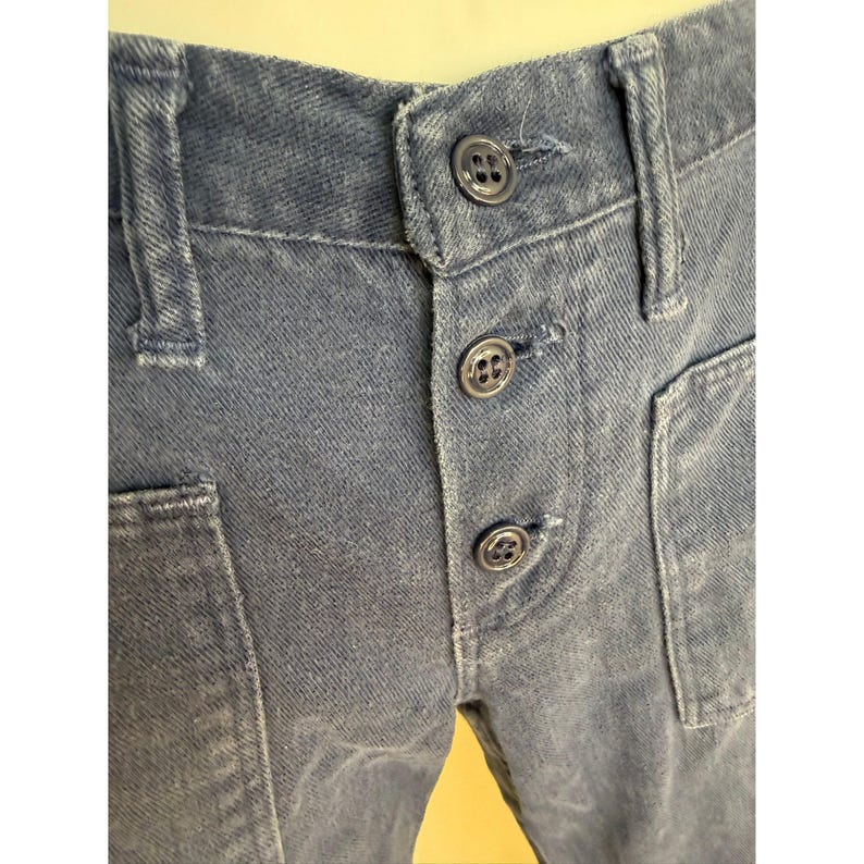 May include: Close-up of blue denim pants with a button fly and two front pockets. The pants have a faded appearance and visible stitching details. The buttons are dark and round.