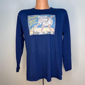 Pode incluir: Uma camisa azul-marinho de manga comprida com uma estampa gráfica retangular de um palhaço em um traje branco com babados e leggings listradas de vermelho e branco. O texto diz Barnum and Bailey Greatest Show on Earth em letras amarelas.