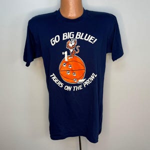 May include: Navy blue t-shirt with white text and a basketball graphic. The text reads "GO BIG BLUE!" and "TIGERS ON THE PROWL". The graphic features a tiger and basketball.