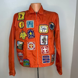 May include: Bright orange vintage jacket adorned with numerous patches. Patches feature text including "Caveman", "Memphis Trail", and "Shiloh Military". The jacket has a zip closure and a collared neckline.