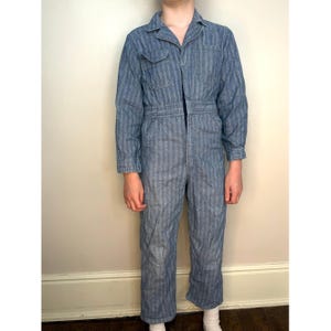 Puede incluir: Mono a rayas azules y blancas con mangas largas, cuello y cremallera frontal. El mono tiene dos bolsillos en el pecho y dos bolsillos en las caderas. La tela parece ser una mezcla ligera de mezclilla o algodón. La prenda la lleva una persona.