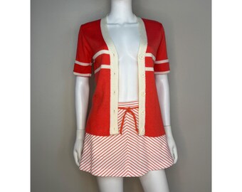 May include: A short-sleeved red cardigan with a cream-colored button-down placket and horizontal cream stripes on the sleeves and chest, paired with a matching red and white chevron-striped skirt featuring a drawstring waist tie.