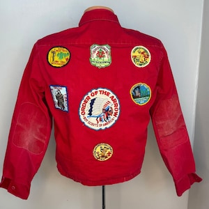 May include: A vintage red jacket with several embroidered patches. The patches display various designs, including text and illustrations related to scouting and camping. The jacket has elbow patches and a button-down front.