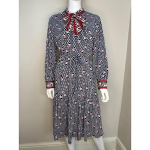 May include: A navy blue dress with a white polka dot pattern and red accents. The dress features long sleeves, a tie waist, and a red bow at the collar. The cuffs and collar have red trim.