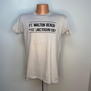 May include: A light beige t-shirt with the text "FT. WALTON BEACH WHERE THE ACTION IS!" printed in black. The shirt is short-sleeved and has a crew neck.